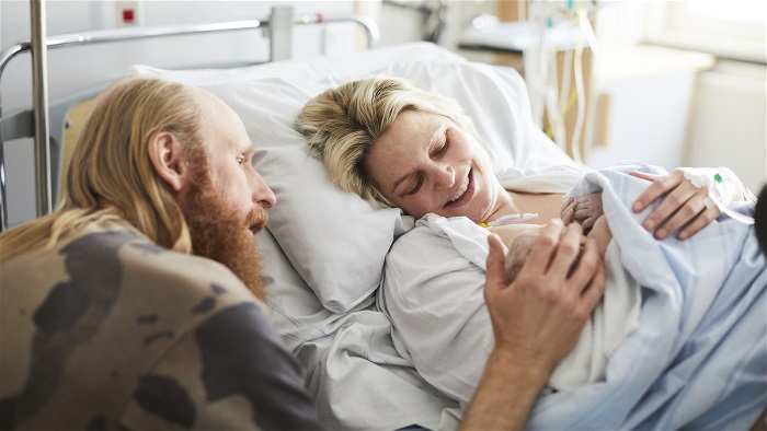 En kvinna ligger i sjukhussäng med nyfött barn och en man sitter bredvid sängen och lägger en hand på bebisens huvud. Foto.