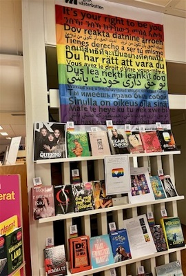 Fotografi med en hylla med böcker och en flagga med texten "Du har rätt att vara du" på olika språk.