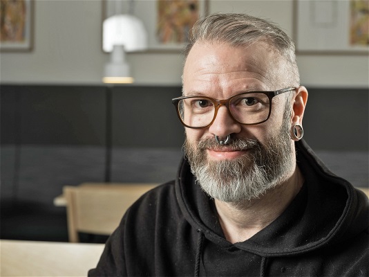 Man med glasögon och piercingar ler och tittar in i kameran.