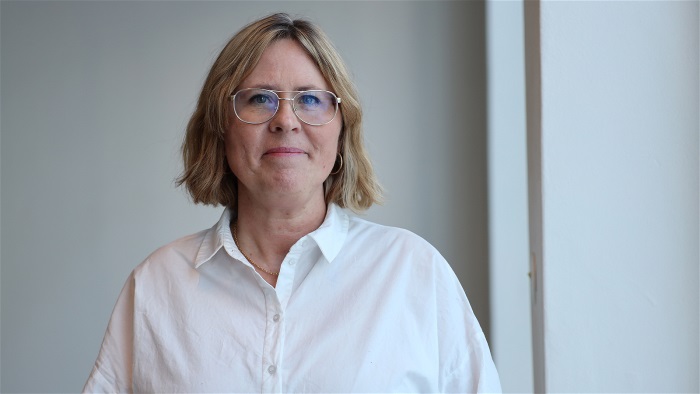 Kvinna med mellanblont hår och glasögon, tittar in i kameran och ler. Foto.