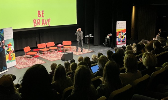 Fotografi som visar en föreläsare på scen och orden "Be brave" skrivet i bakgrunden.