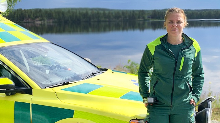 Kvinna i ambulansklädsel står framför ambulans vid vattnet. Ser allvarlig ut. Foto.